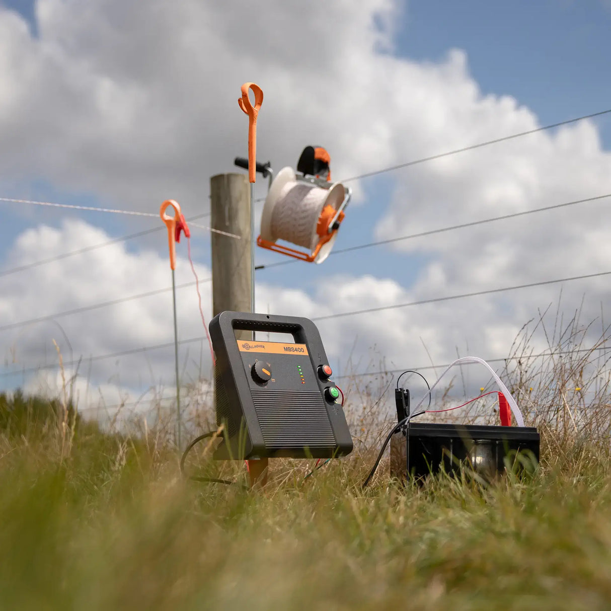 Electric fence energizer and components on a grassy field with a cloudy sky.
