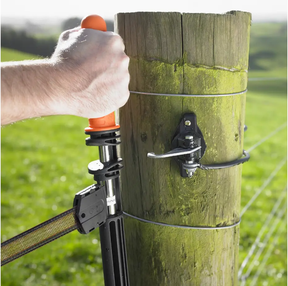 Hand using a fence post connector tool on a wooden fence post with a grassy field background.