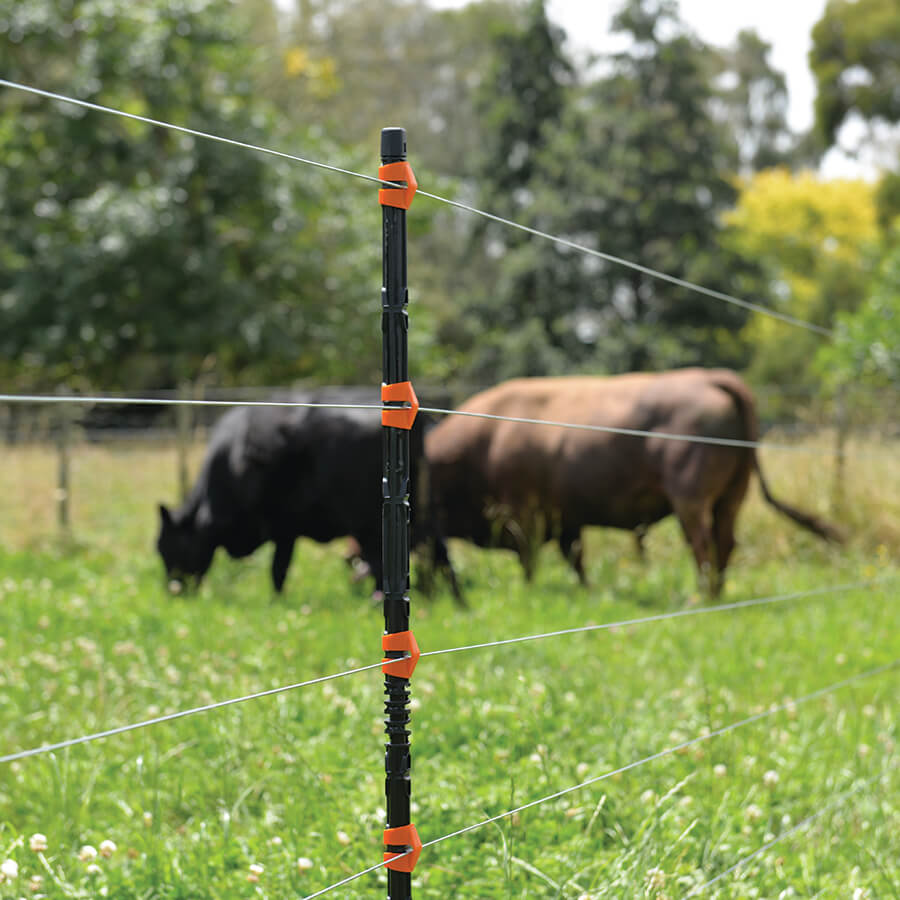 An insulated line post for electric fencing, with orange and black coloring, installed in a field with a cow in the background.