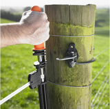 Person using a fence post connector tool on a wooden fence post with a grassy field background.
